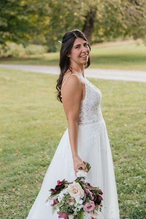 Radiant bride in lace gown smiles over her shoulder while holding a burgundy and blush bouquet outdoors.