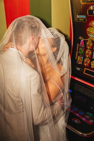 Bride and groom kissing under veil beside colorful casino slot machine at wedding reception.