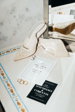 Wedding flat lay featuring pearl-studded bridal heels, gold rings, and elegant invitation suite on marble surface.