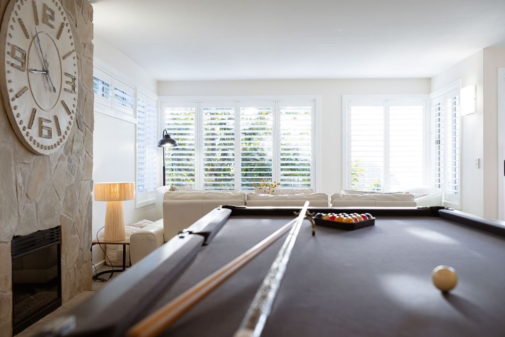 Modern game room with pool table near bright windows and large wall clock in contemporary white interior space.