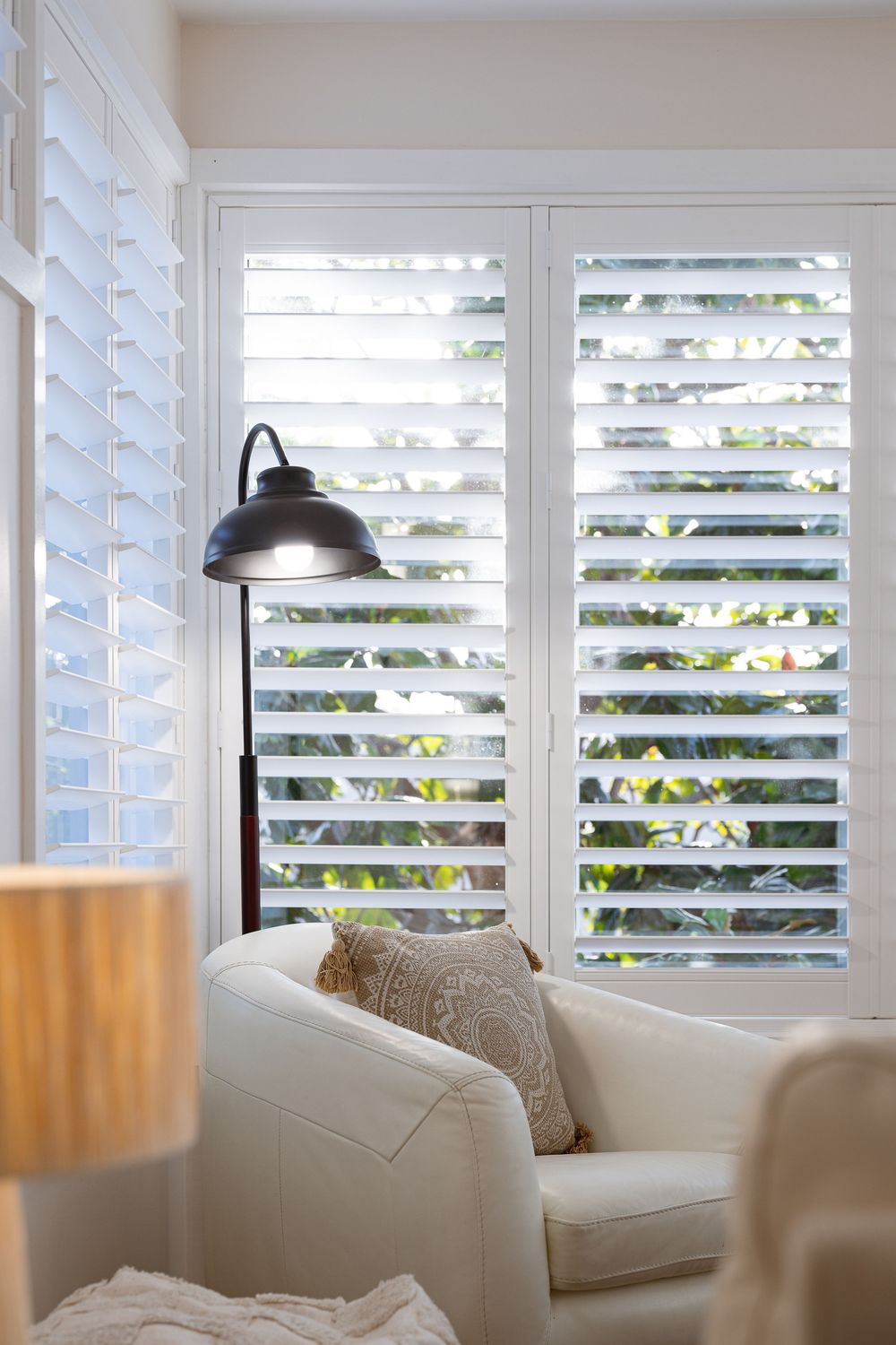 White plantation shutters filter natural light into a bright living room with cream-colored upholstered furniture.
