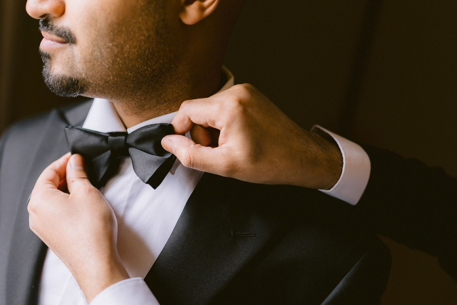 A person adjusts a black bow tie while wearing a formal tuxedo in an elegant setting.