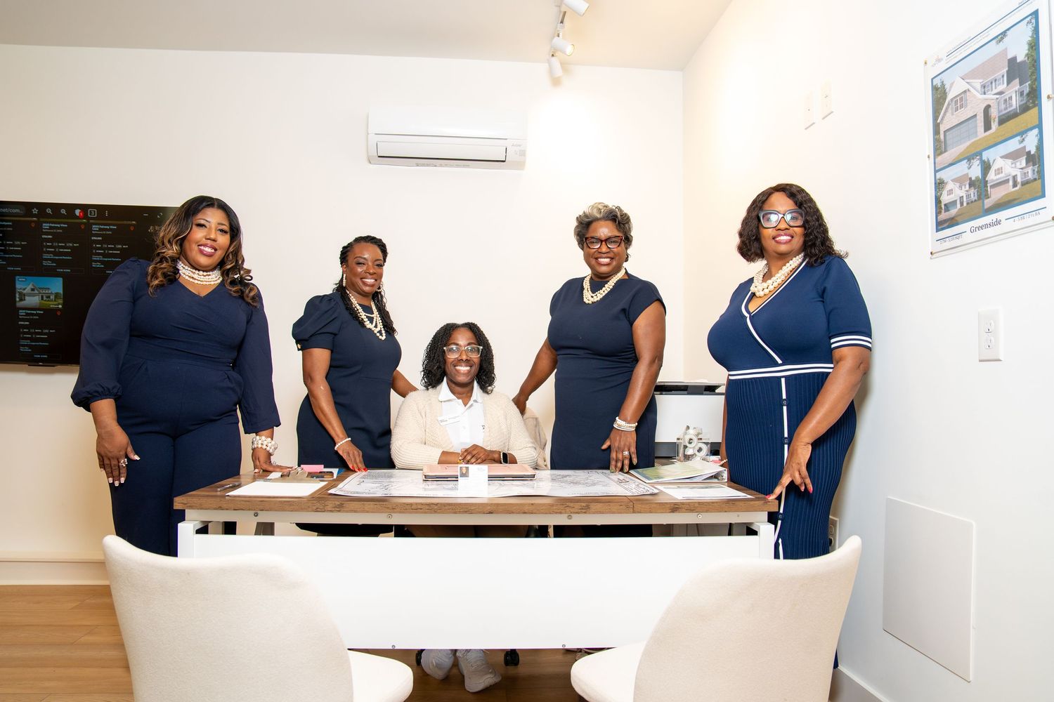 Five women in navy outfits gather around a desk in a real estate office, smiling as they pose together.