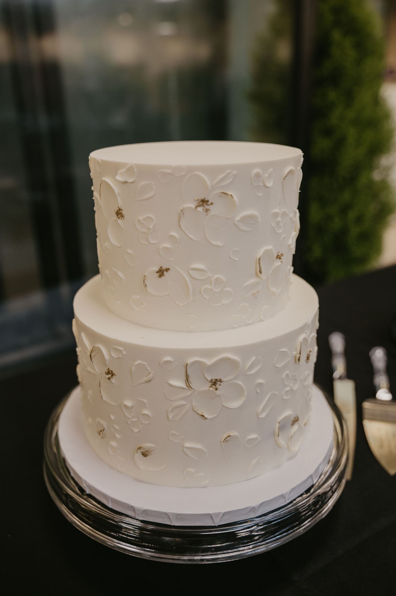 Two-tiered white wedding cake with delicate floral embossing and pearl details on a silver cake stand.