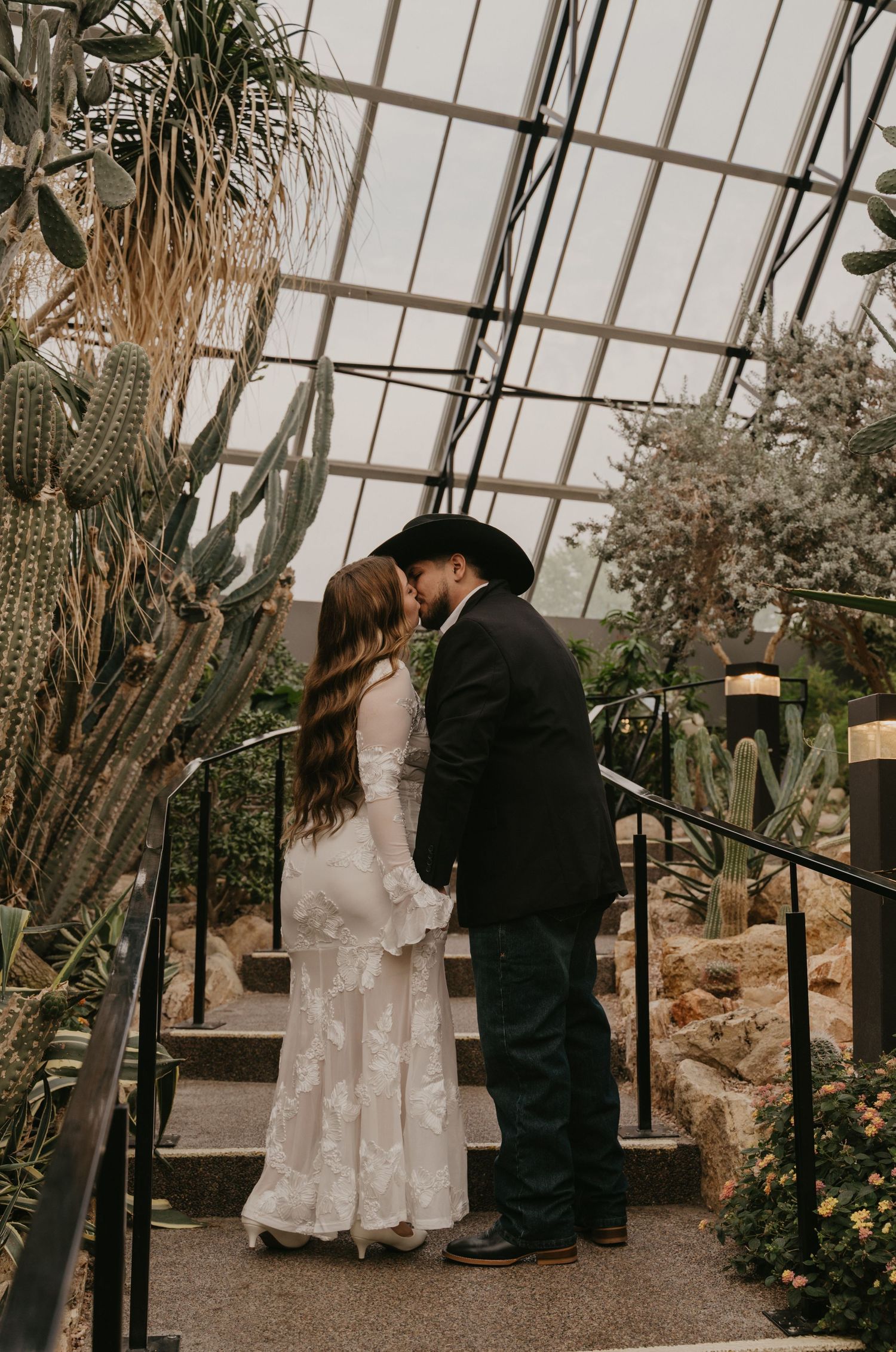 A romantic wedding couple shares a kiss in a greenhouse filled with cacti and tropical plants.