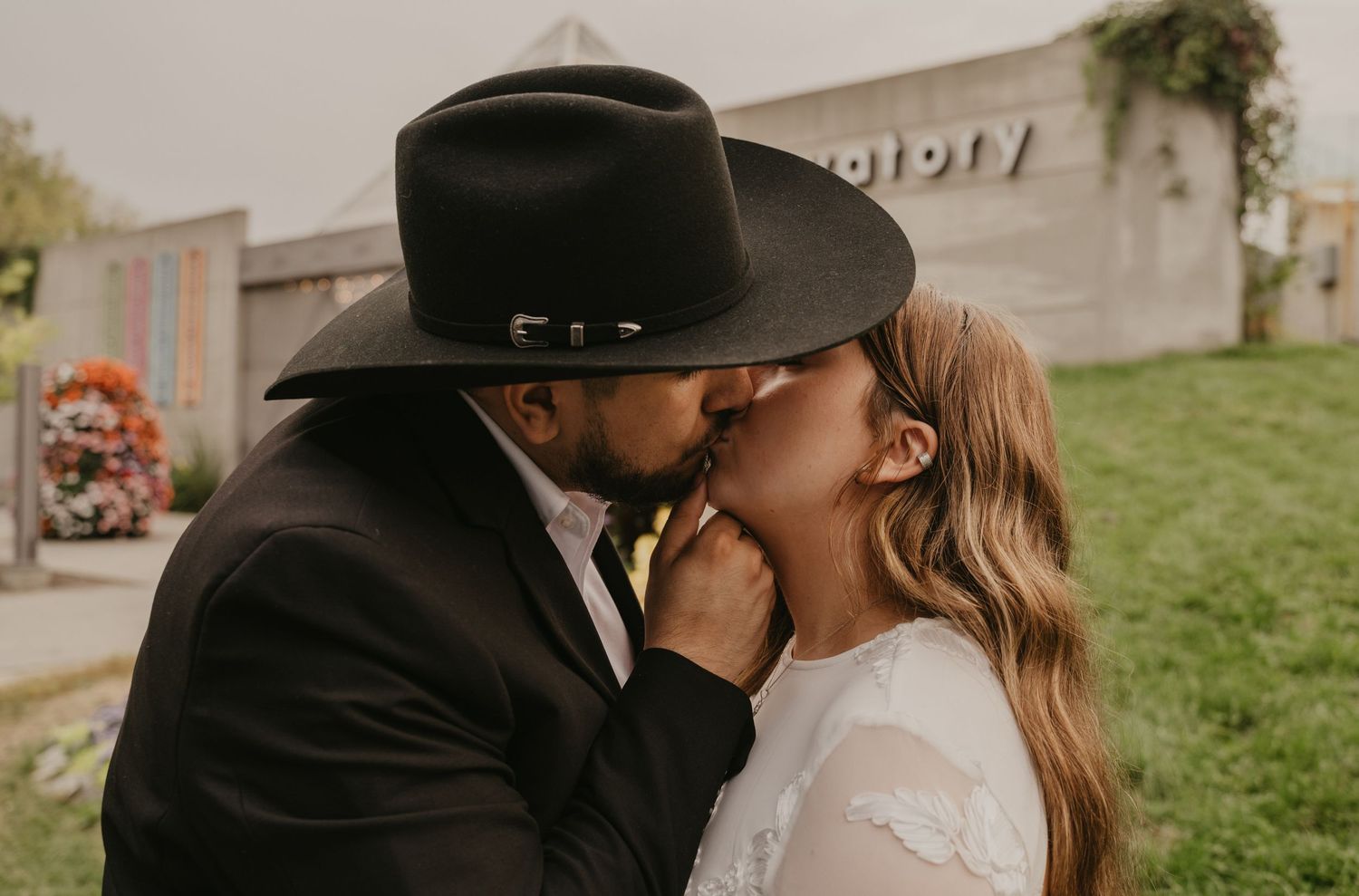 A romantic wedding moment captured outdoors with modern architecture and greenery in the background.