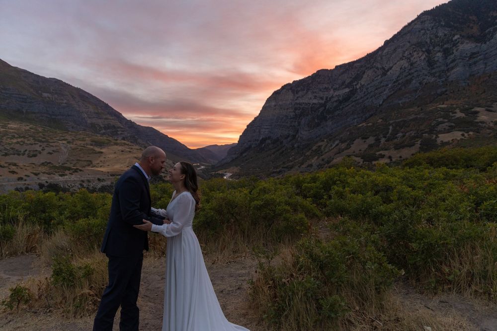 A couple in wedding attire stands silhouetted against a dramatic mountain sunset in a scenic outdoor landscape.