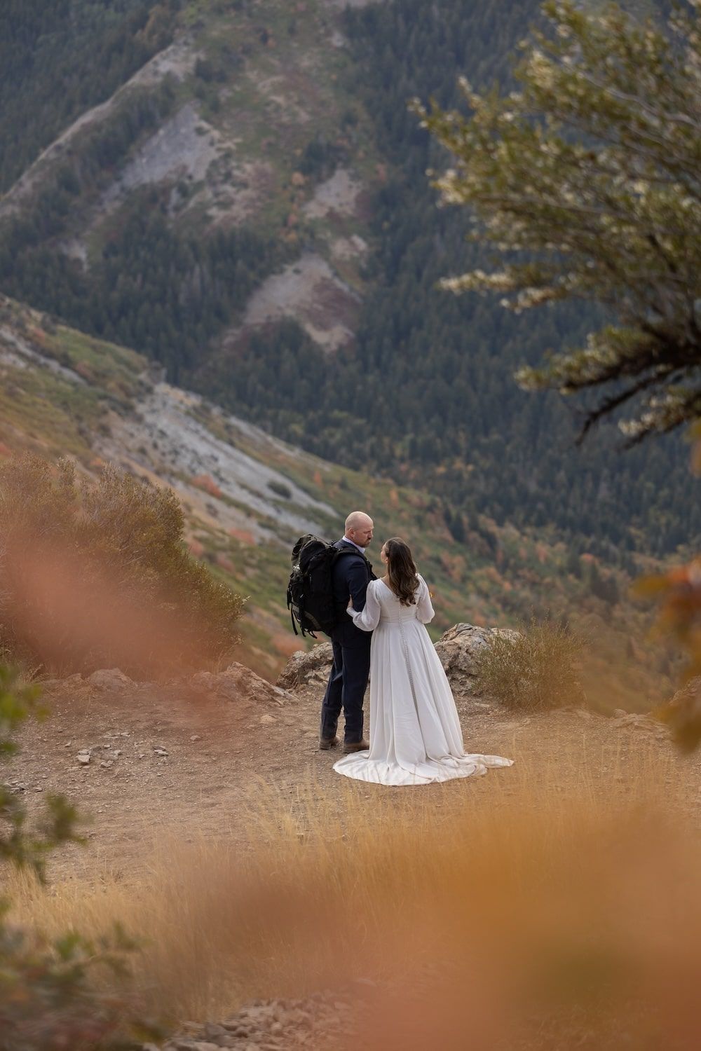 Two figures stand together on a mountain overlook surrounded by autumn grasses and rugged peaks.