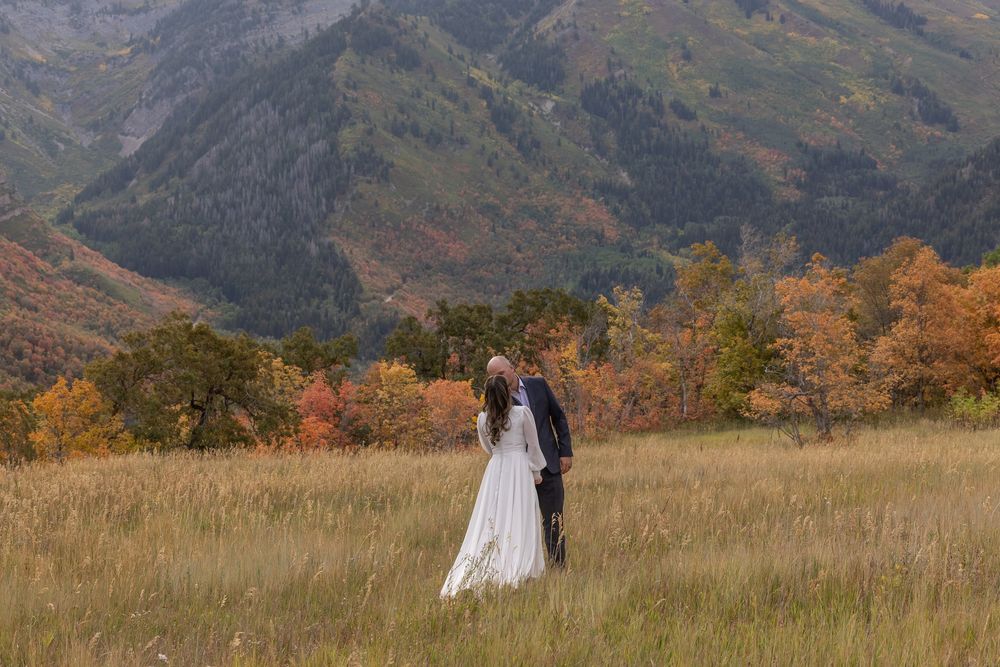 A couple in wedding attire stands together in a scenic mountain meadow with autumn foliage and peaks in the background.