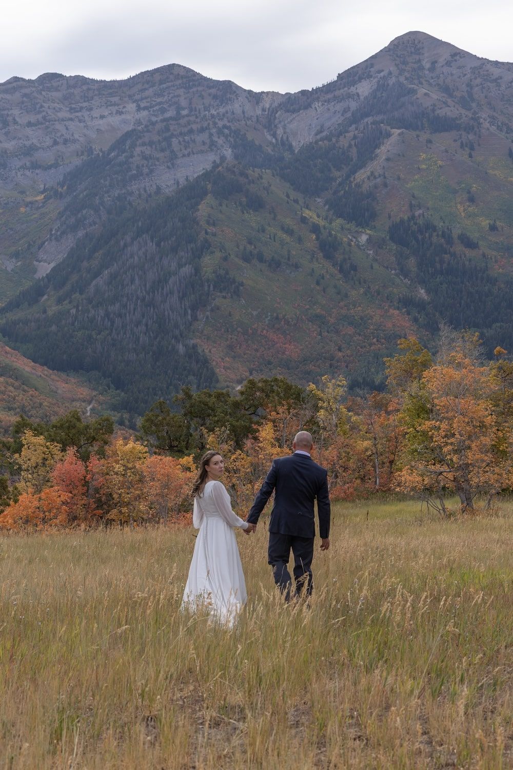 A couple walks through tall grass with majestic mountain peaks and fall foliage in the background.