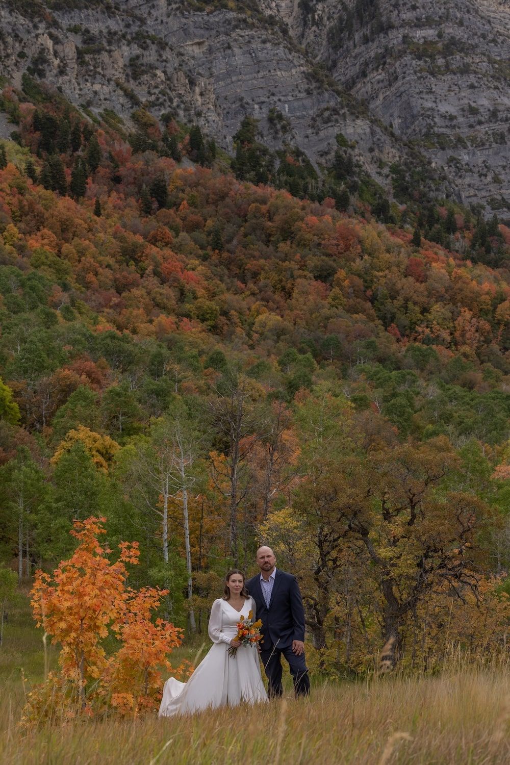 Wedding couple walks through golden fall foliage on mountainside with dramatic cliffs in background.