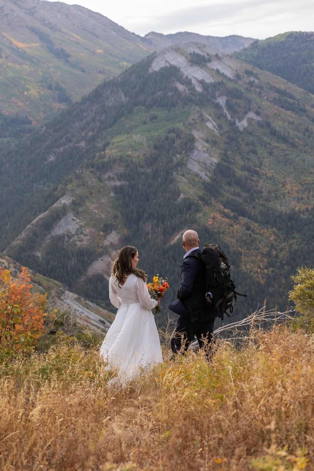 A couple in wedding attire sits on a mountain overlook surrounded by autumn foliage and dramatic mountain vistas.