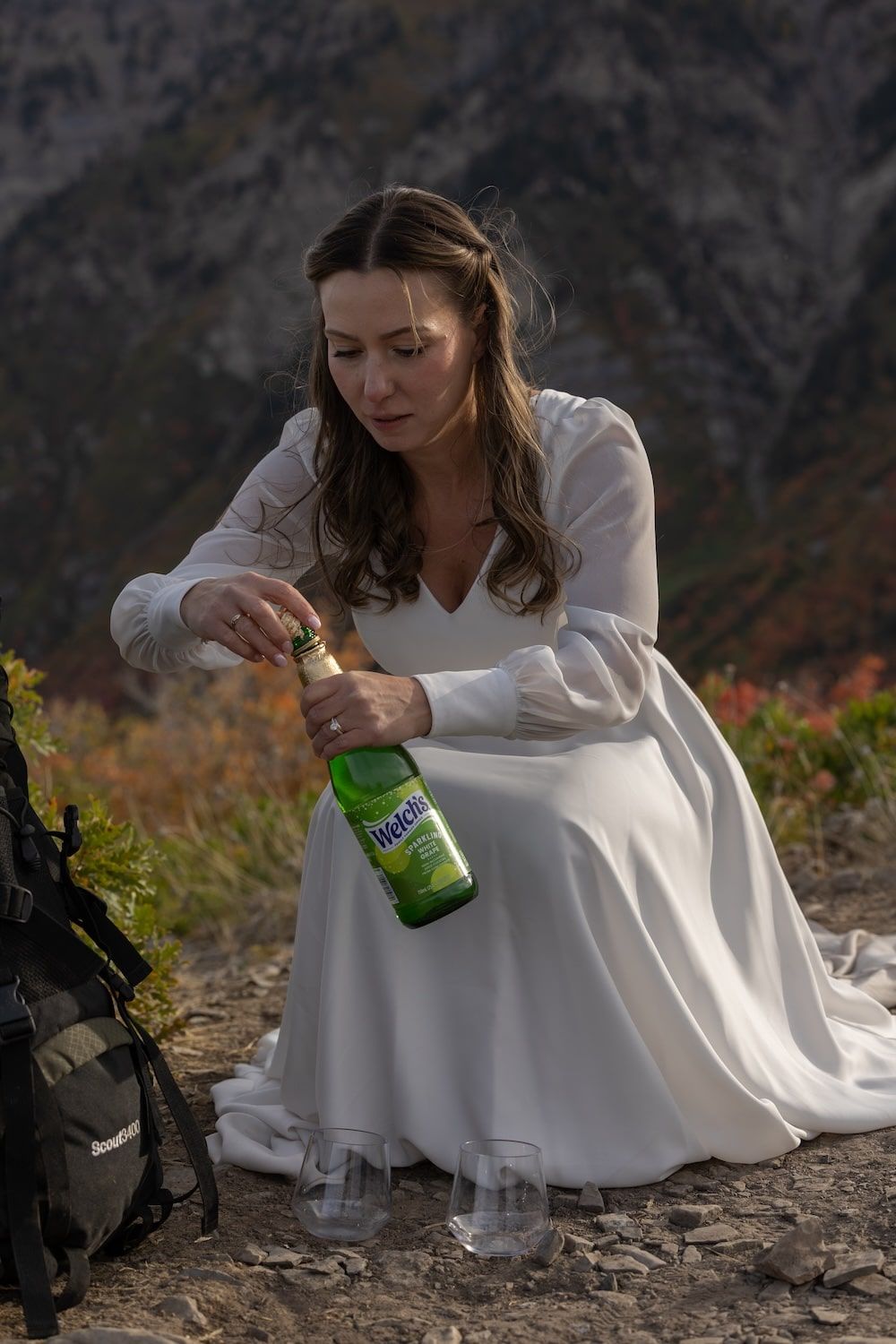 A person in a white dress pours champagne while sitting on a rocky mountain ledge with dramatic cliffs in background.