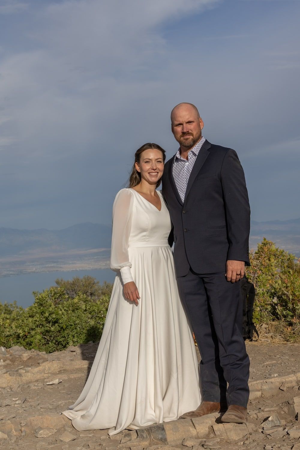 A couple poses together outdoors on a rocky overlook during a wedding photoshoot against a blue sky.