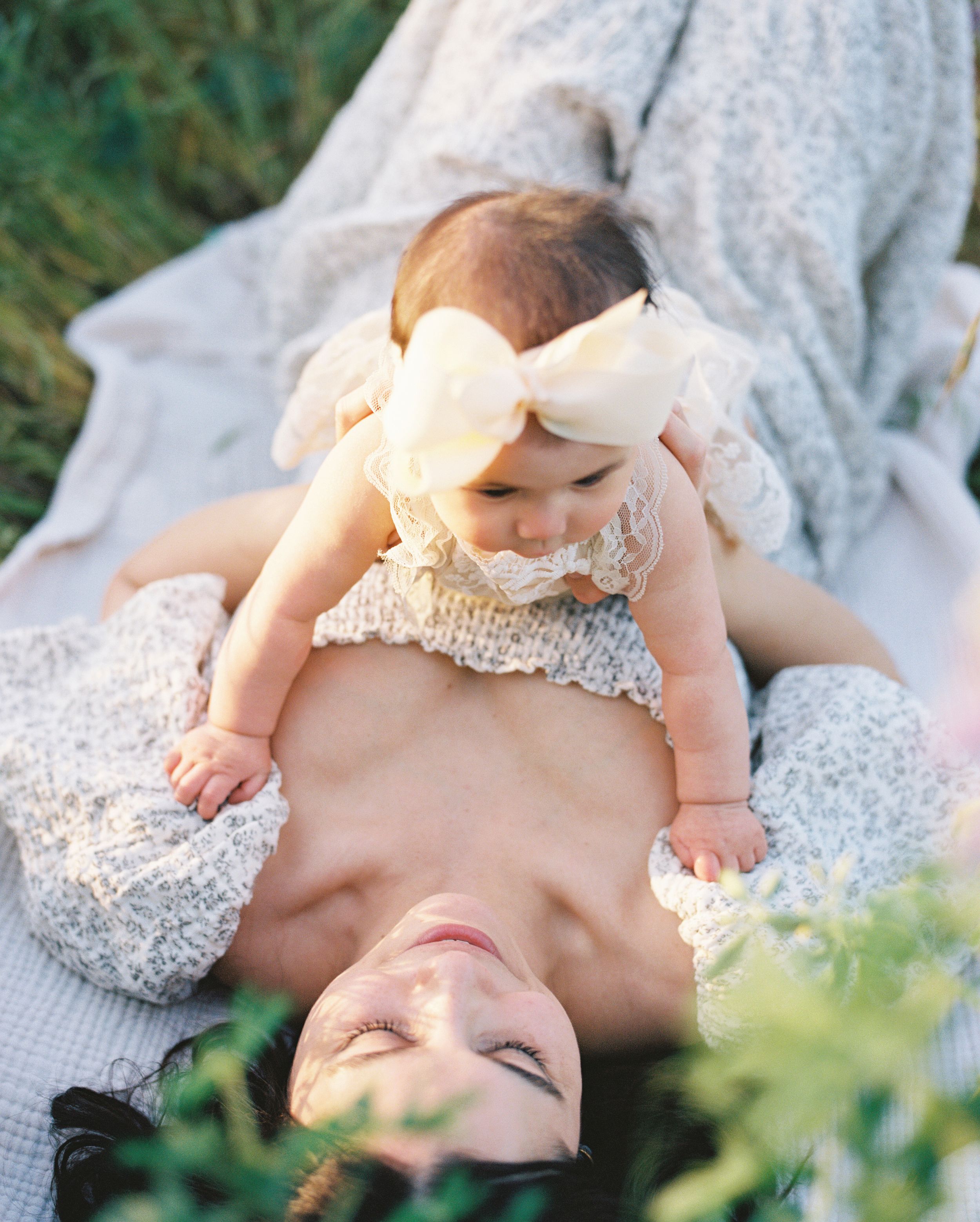 A baby wearing a bow lies on a blanket outdoors in summer.