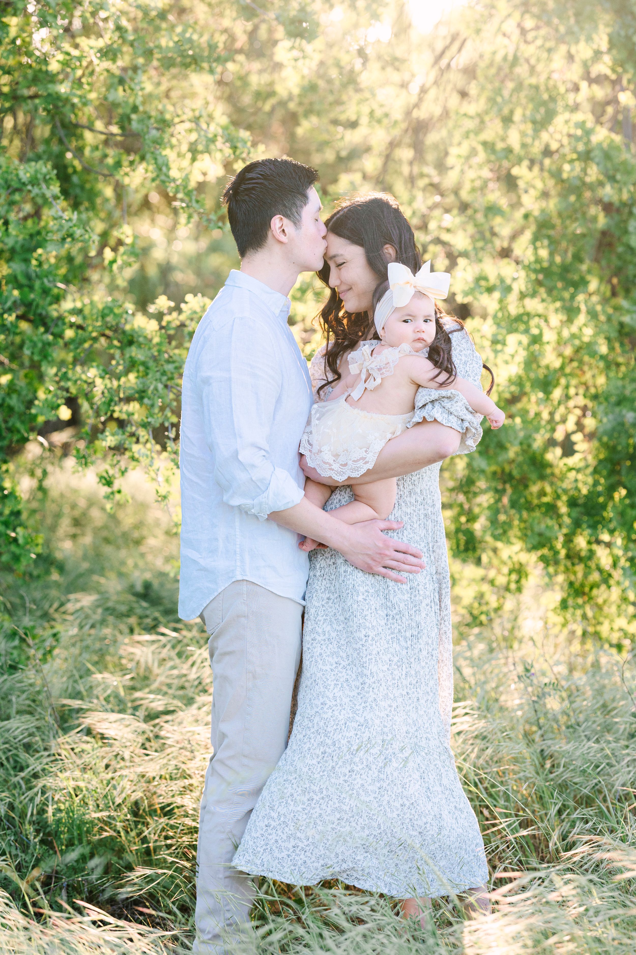 A family in white and beige clothing embraces in a sun-dappled outdoor setting.