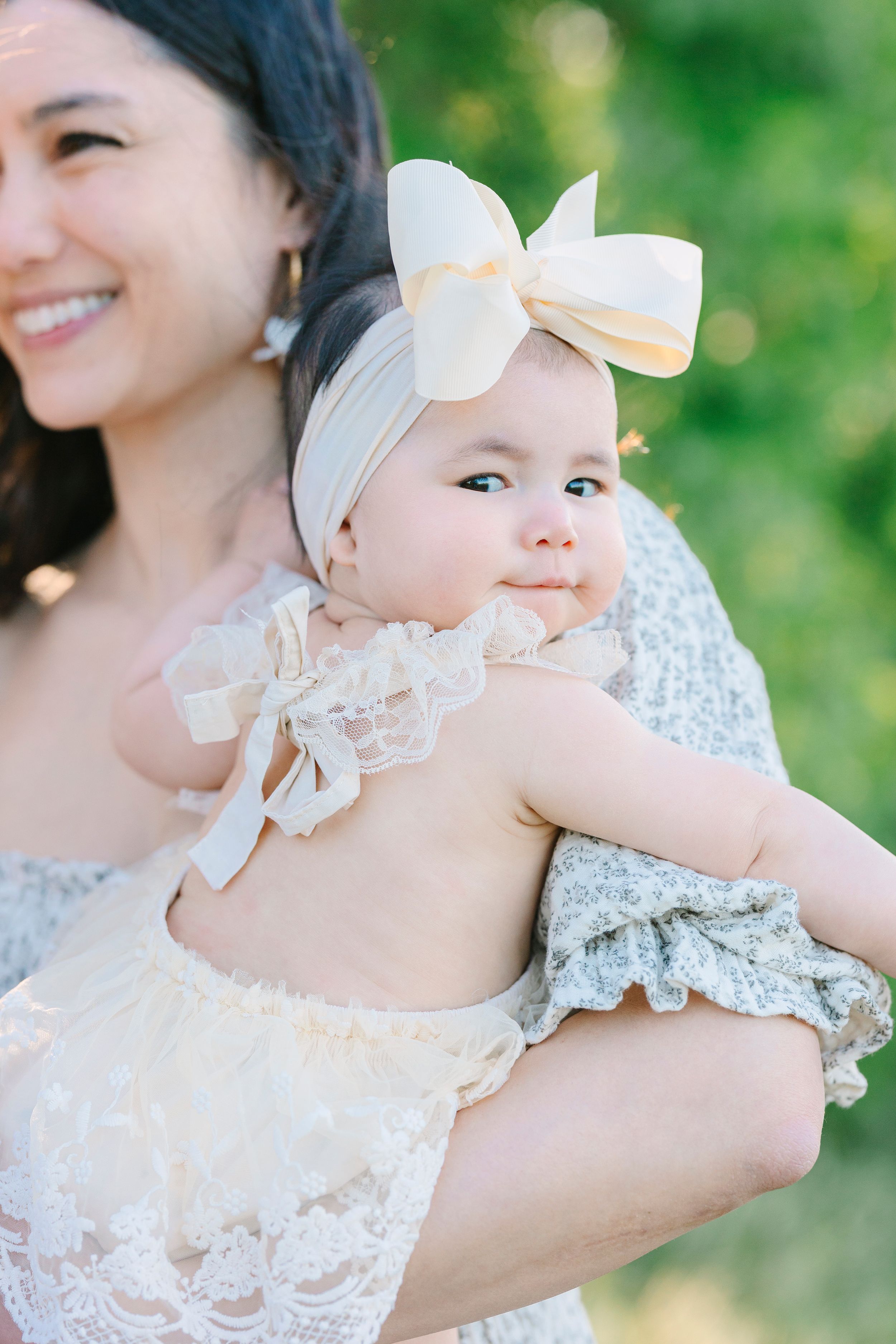 A baby wearing a white bow and lace dress is held lovingly outdoors.
