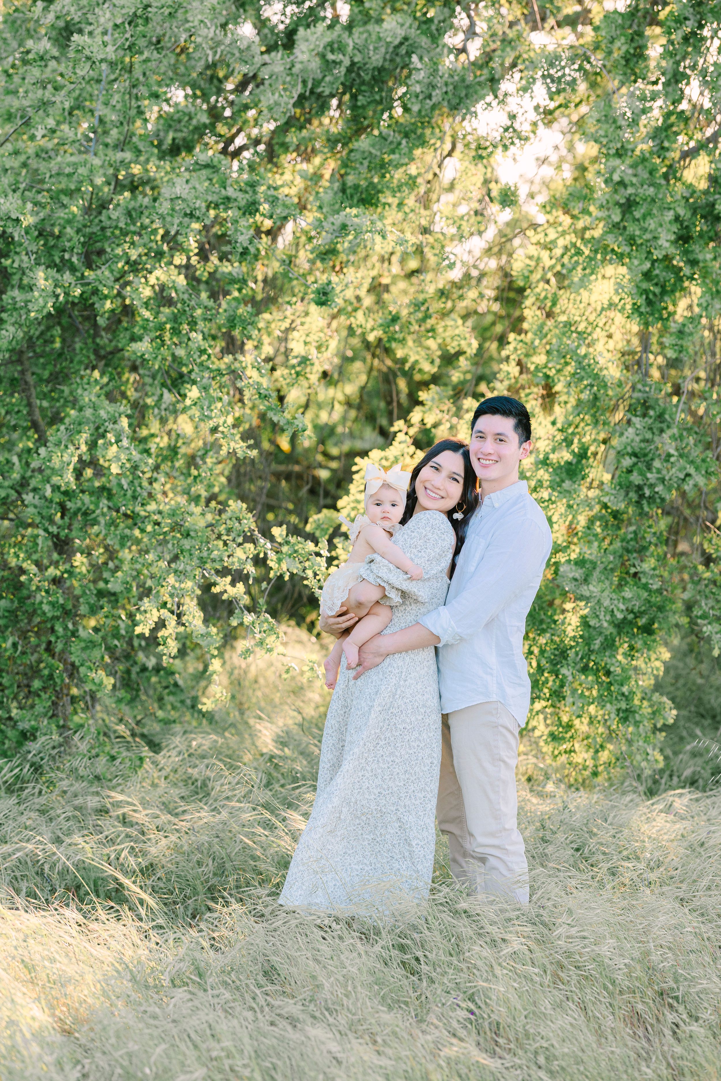 A family in light colored clothing poses together in tall grass with sunlit trees behind them.