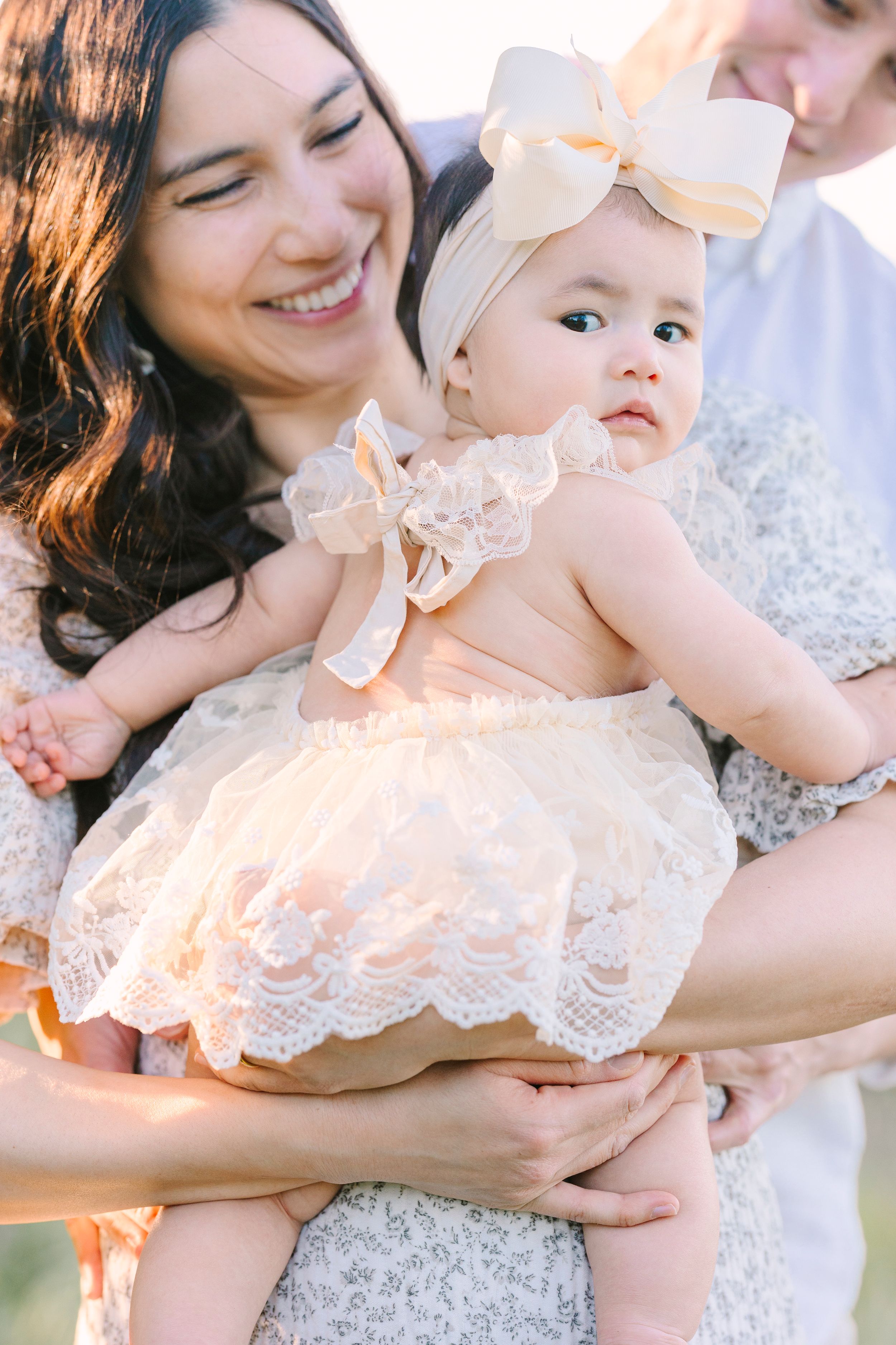 A baby in a lace dress with bow is held lovingly in warm sunset light.