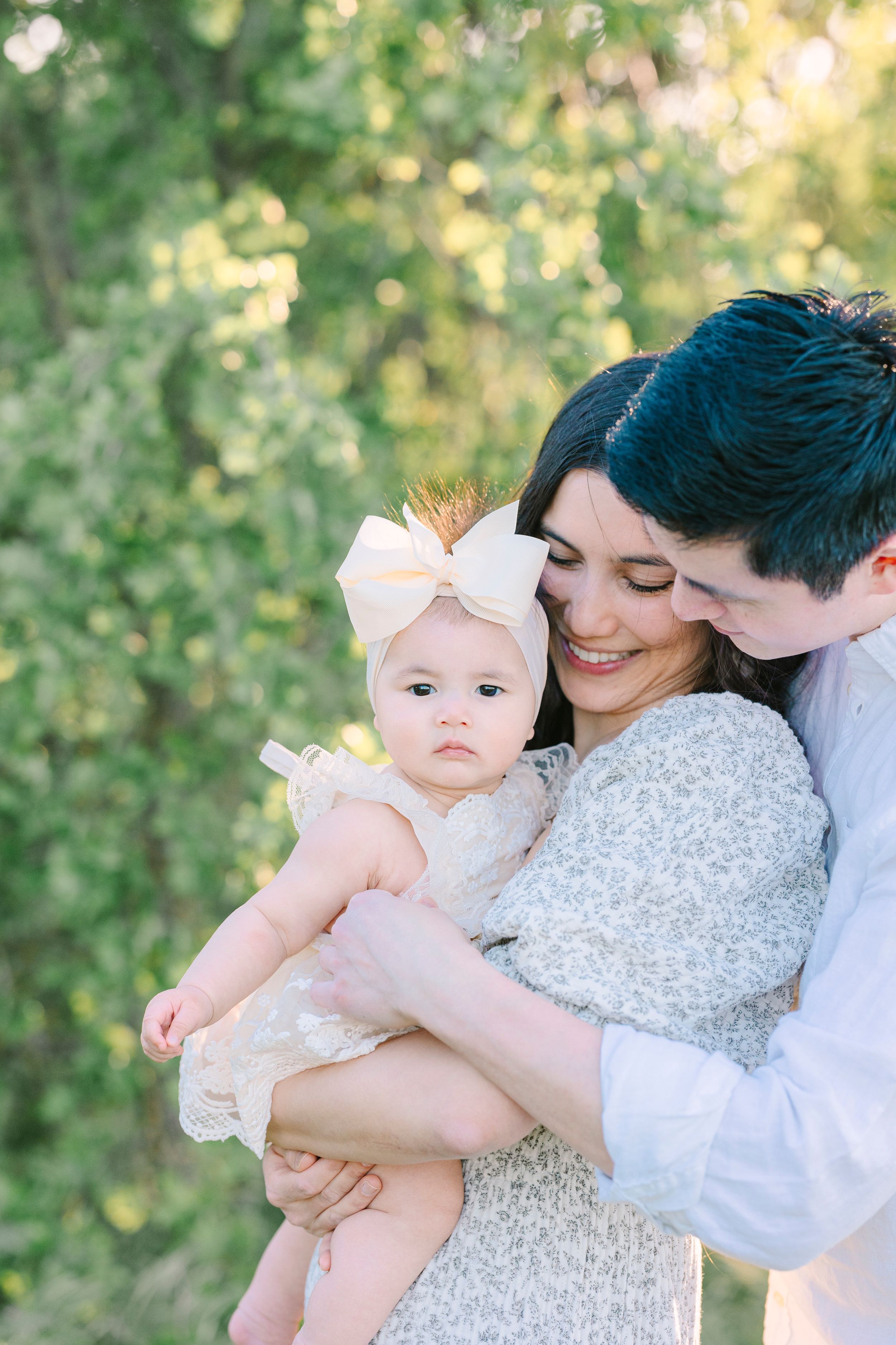 A family shares tender moments with their baby against a backdrop of sunlit greenery.