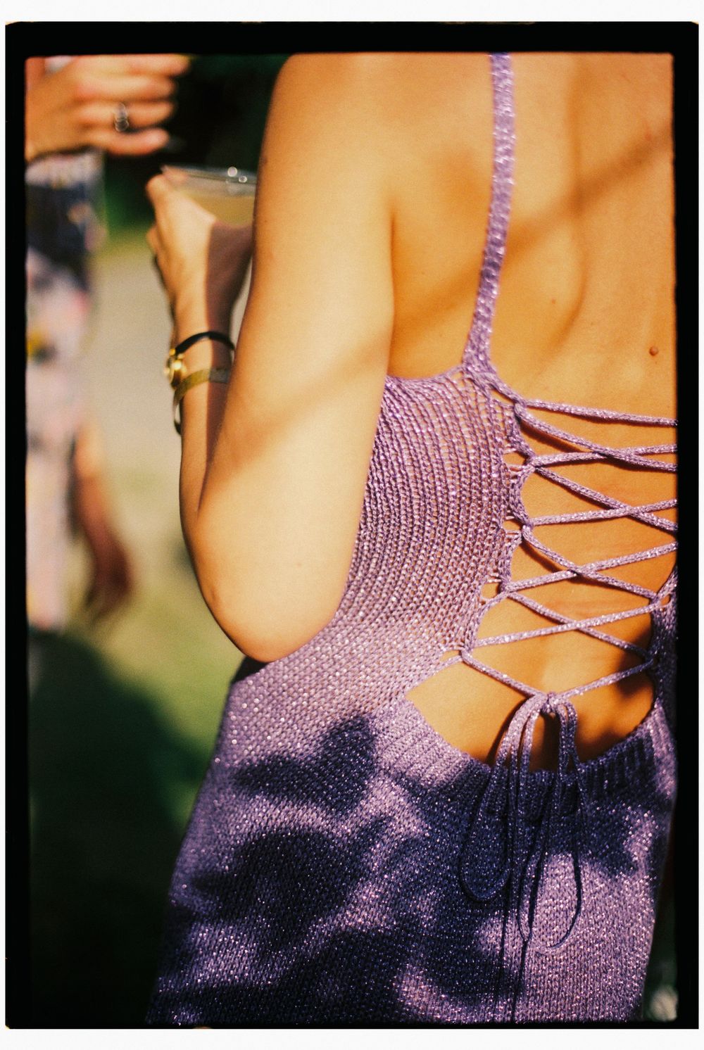 Detail of the sun shining on a guest's purple knit dress with a tie up back while she holds a glass of wine and the leaves create shadows.