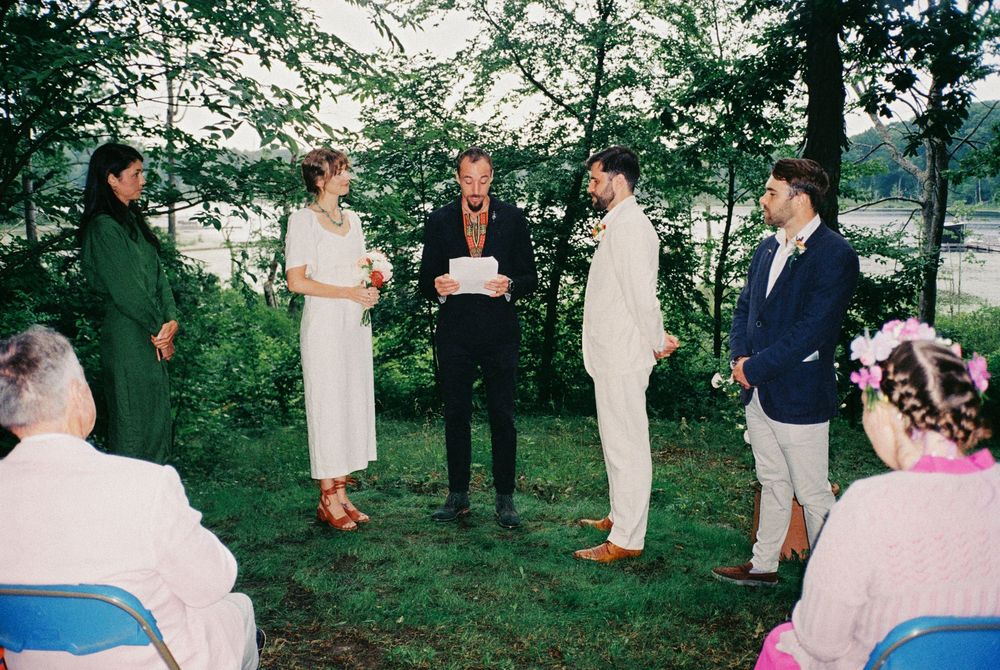 The wedding officiant stands between the couple in front of the trees and lake while guests watch their loved ones say their ceremony vows.