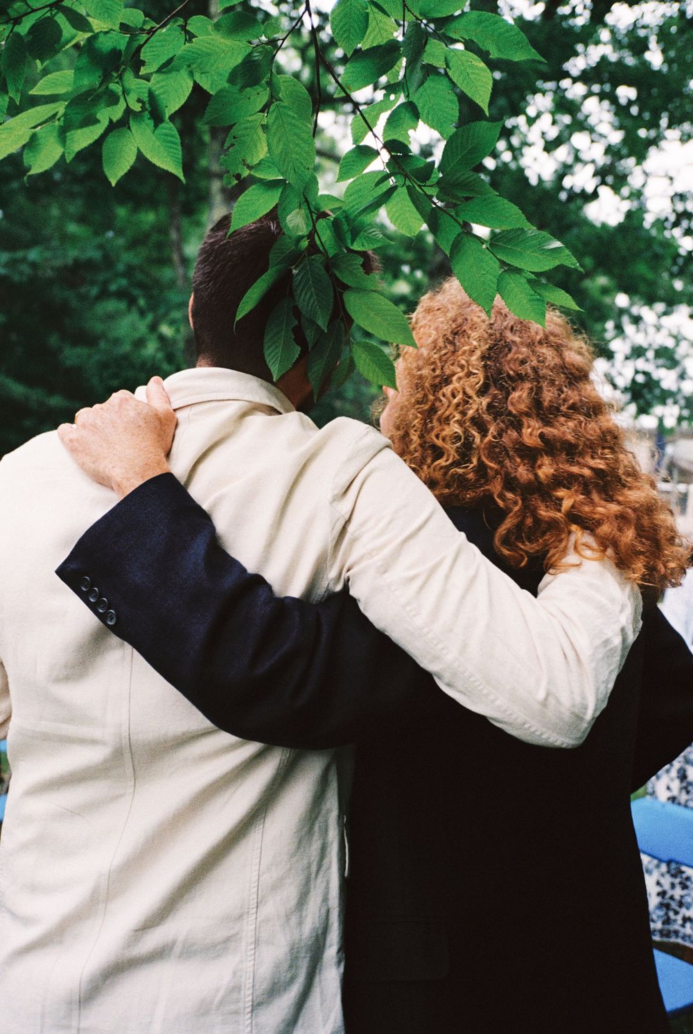 The groom & his cousin with their arms around each other under green tree leaves. One wears a light linen suit and other with long red hair.