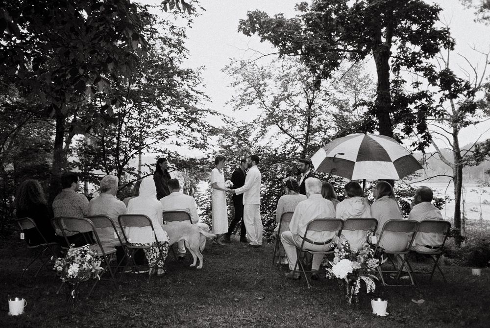 Black and white film exposure during the ceremony showing all the guests holding umbrellas under the trees while the couple holds hands.