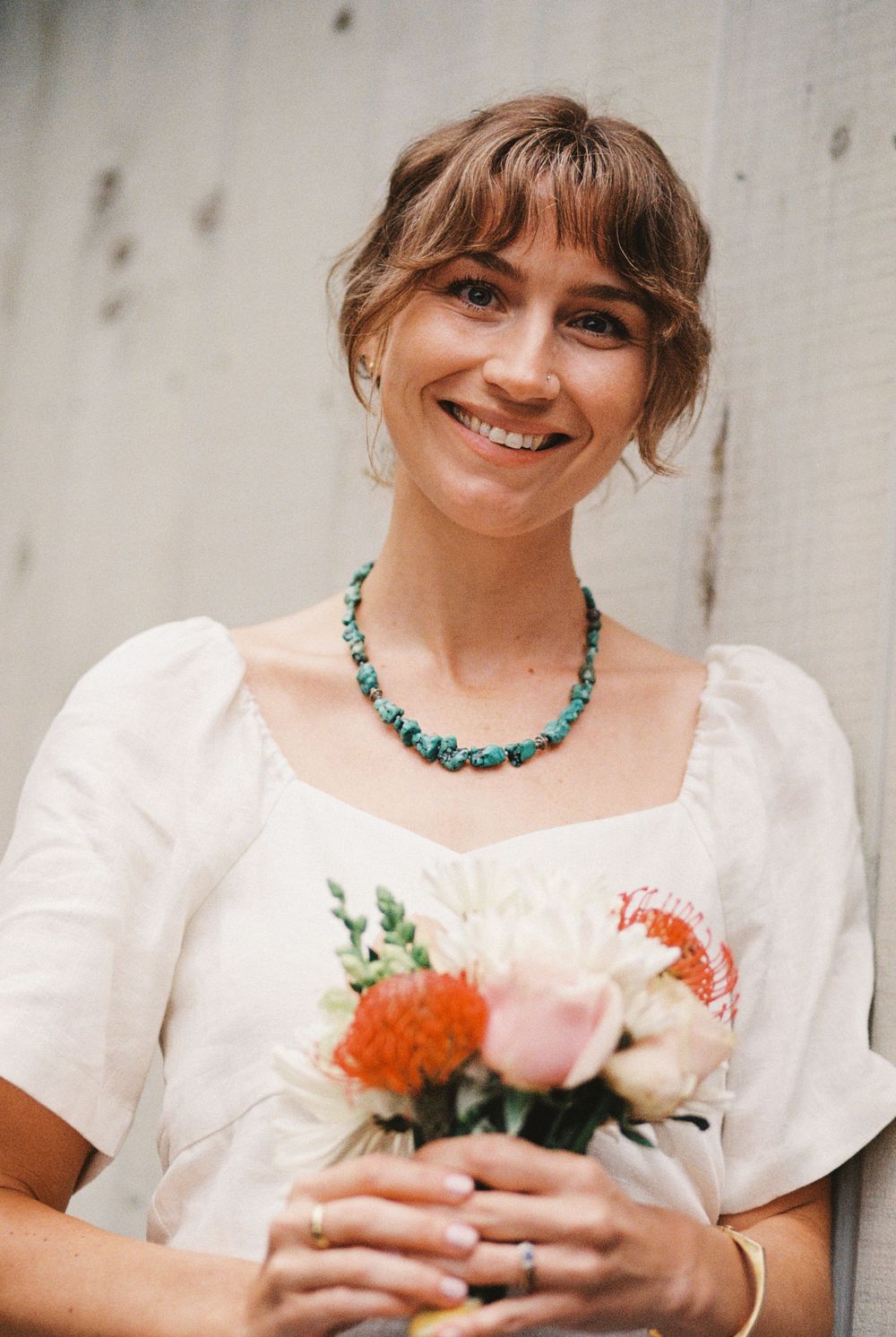 Portrait of the bride holding her colorful bouquet and wearing a turquoise necklace with her linen wedding dress in front of their home.