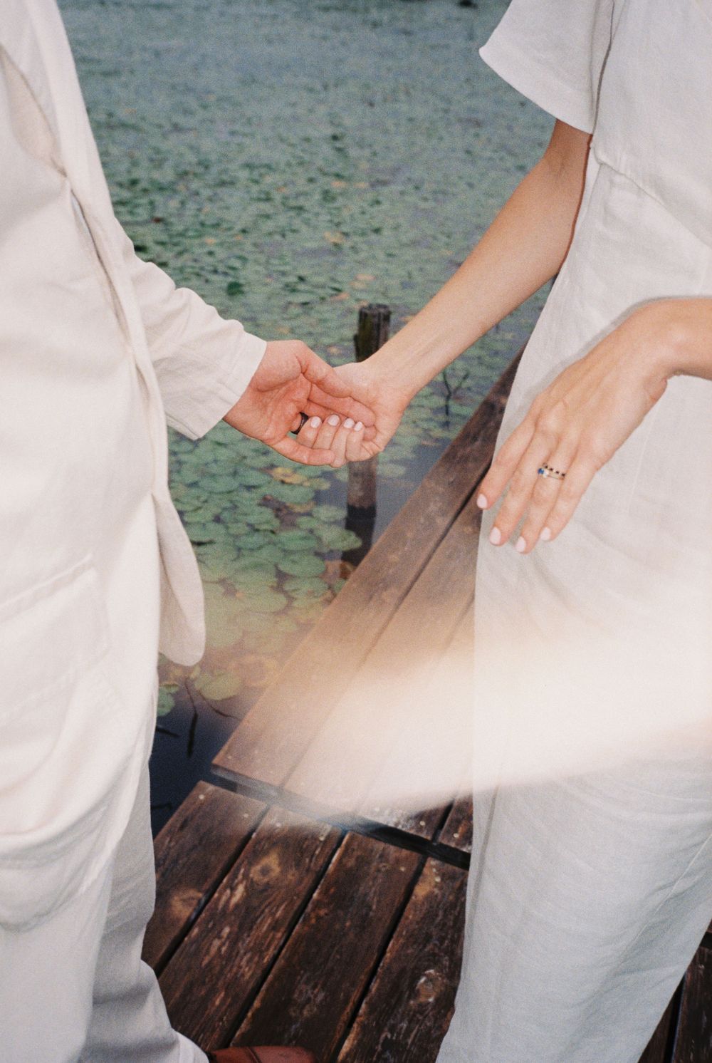 35mm color film image of couple holding hands on a wooden dock, pale green lily pads in the background and a cream light leak on their linen