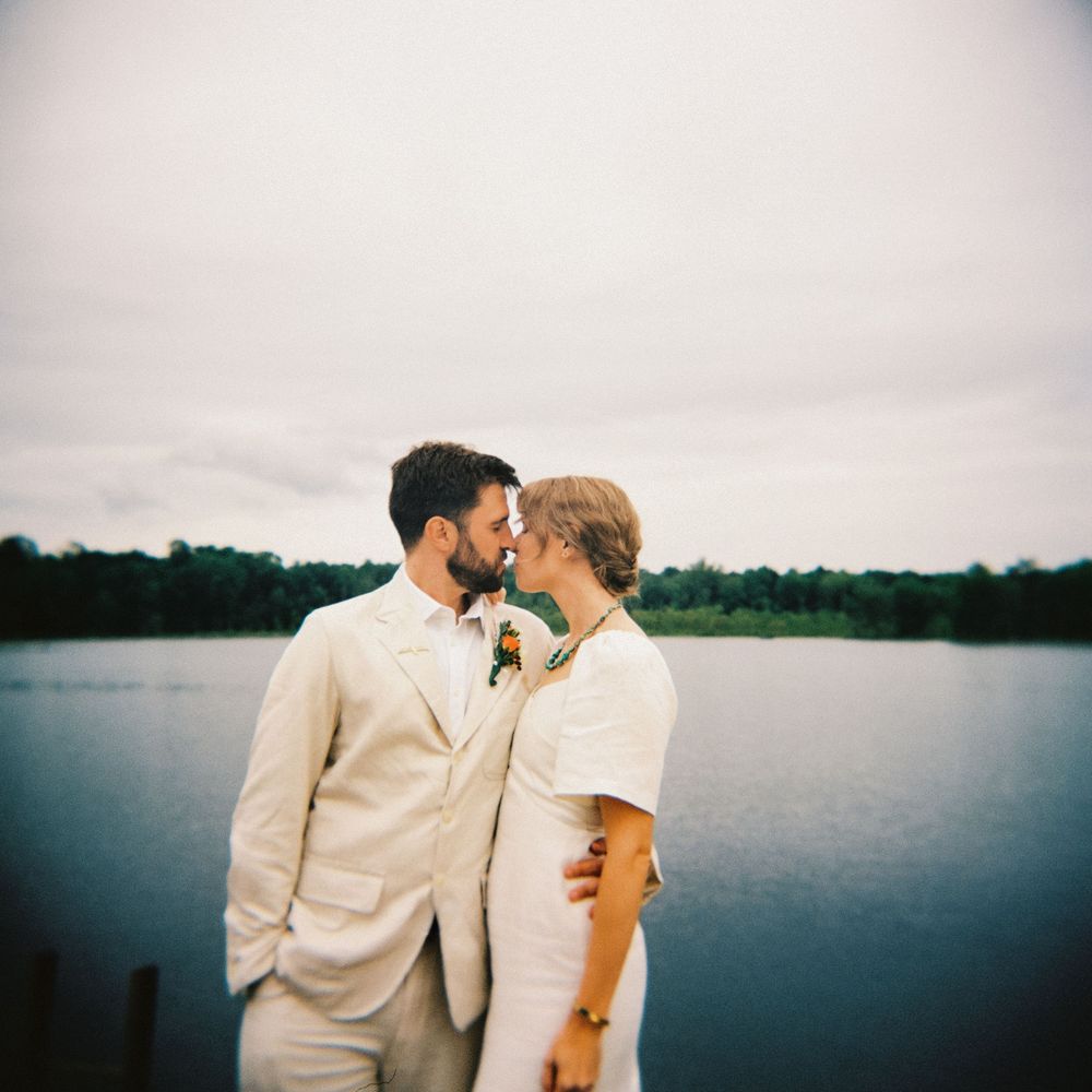 120mm color film photo of the wedding couple, both dressed in cream linens as they lean in to kiss eachother by the lake in Upstate New York