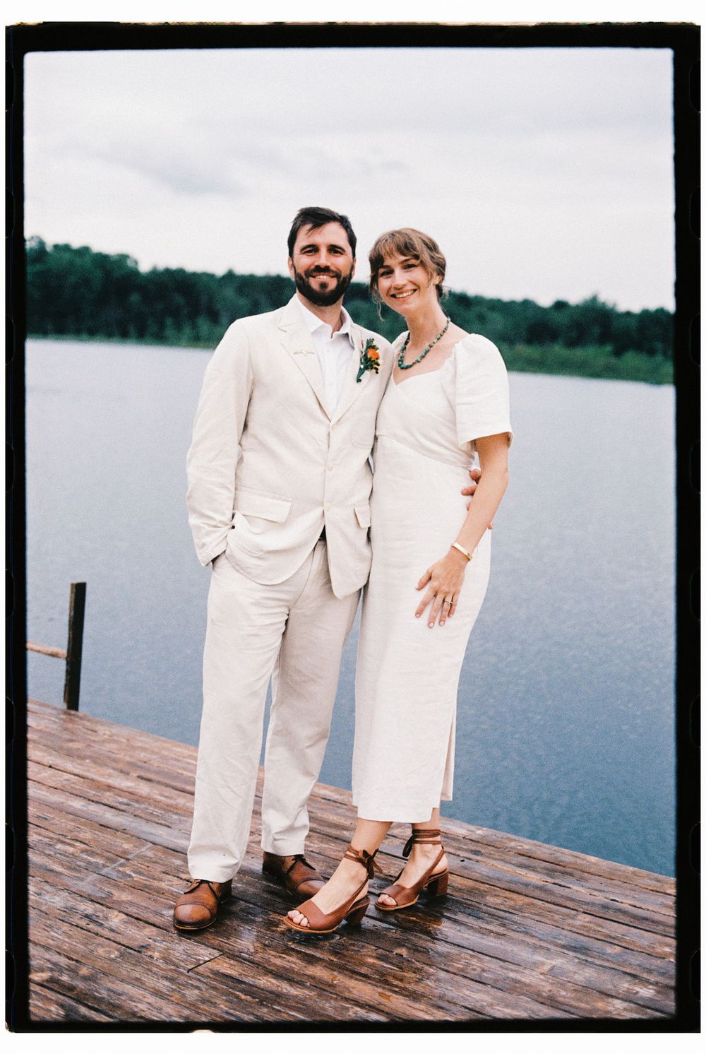 Portrait of the couple after their ceremony on a dock by a pristine lake on 35mm film. The sky is soft and overcast and they are smiling.