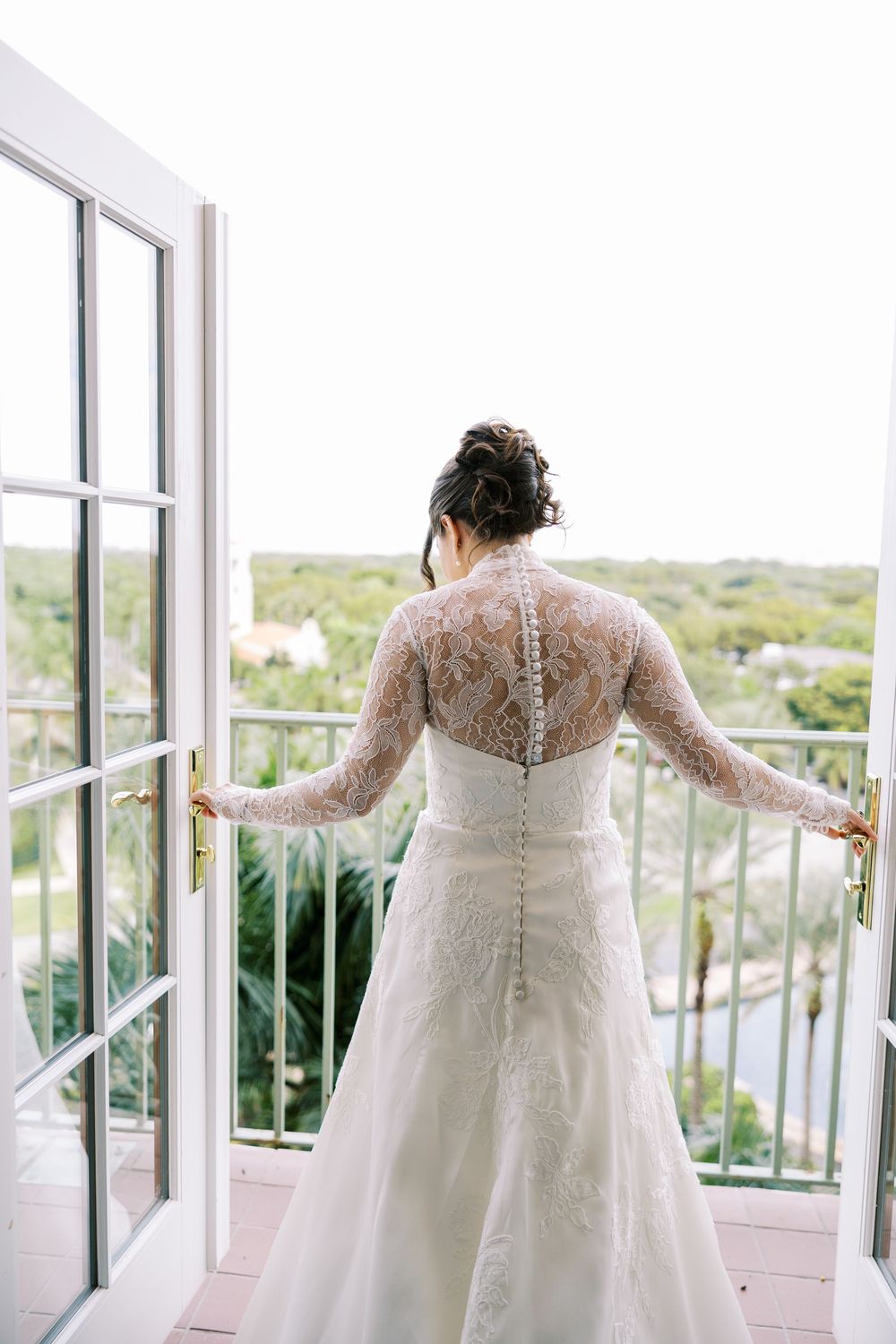 A bride in a lace long-sleeved wedding gown stands by large windows overlooking a scenic view.