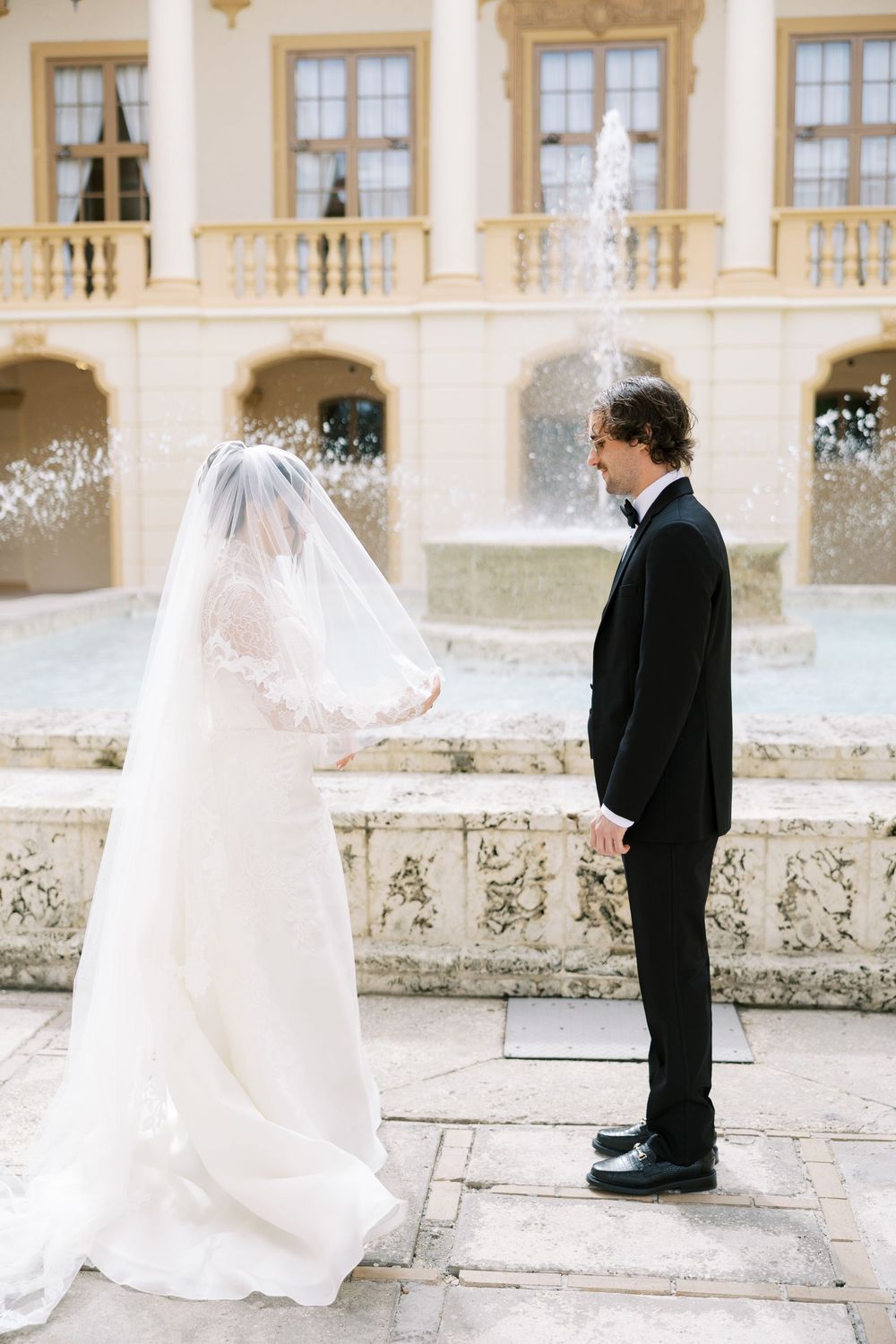 A romantic first look moment between a bride in white and groom in black at an elegant stone courtyard.