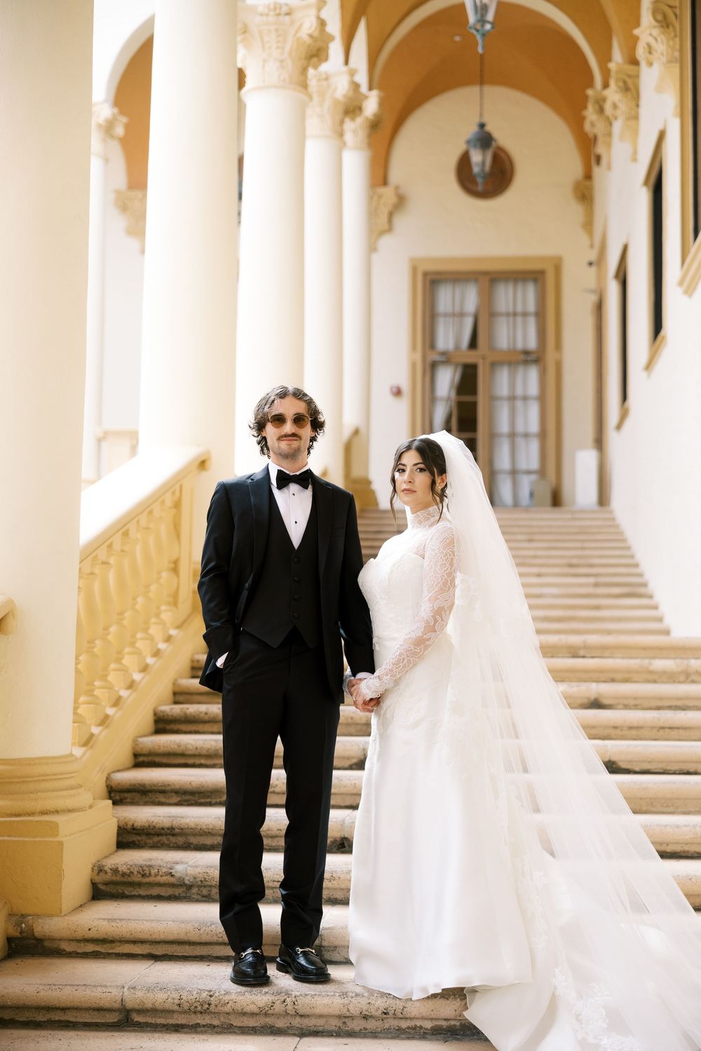 A wedding couple poses on elegant marble steps with ornate columns in a classic architectural setting.