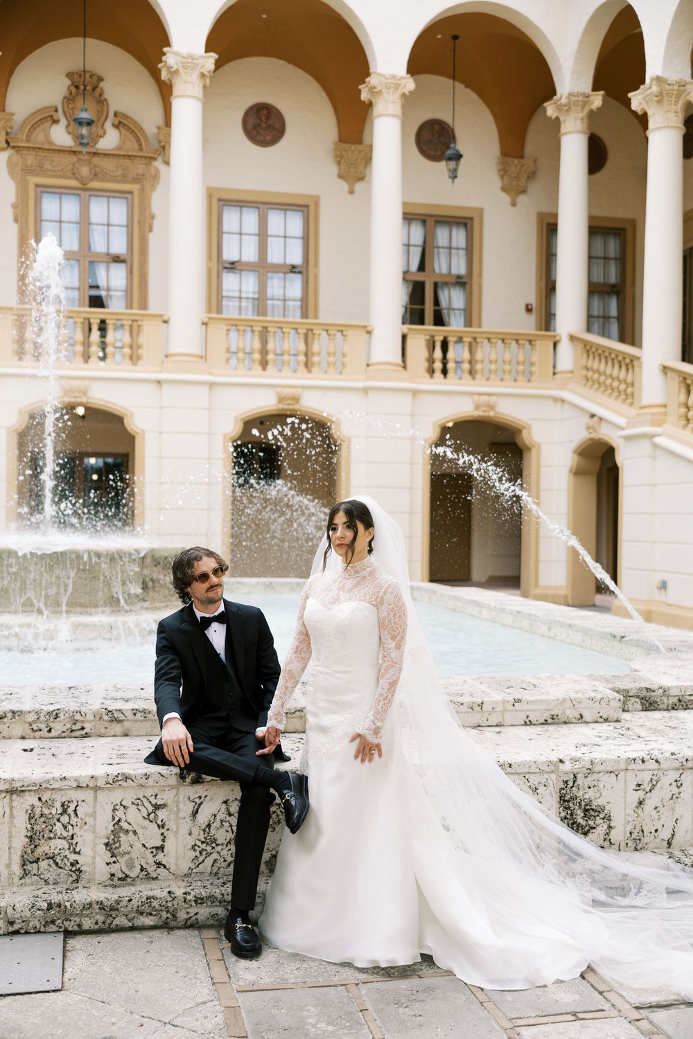 A wedding couple poses by an elegant fountain at a Mediterranean-style venue with arched columns and balconies.