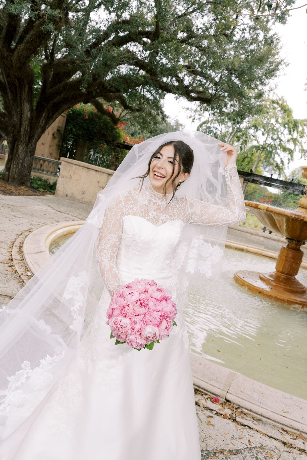 A bride in a white lace gown holds a pink rose bouquet while posing near a garden fountain.