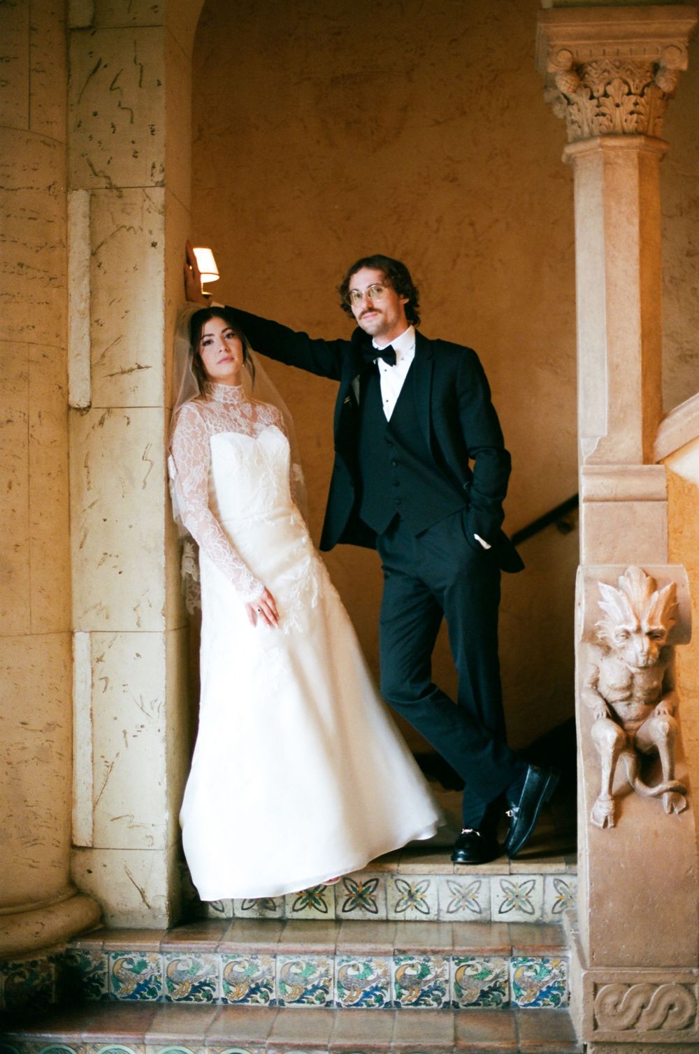 A couple poses on ornate stone steps, with the bride in a white lace gown and the groom in a classic black tuxedo.