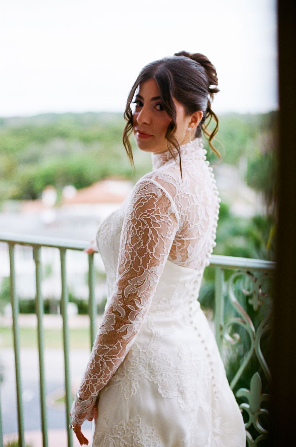 A bride in a long-sleeved lace wedding gown poses elegantly on a balcony overlooking a scenic landscape.