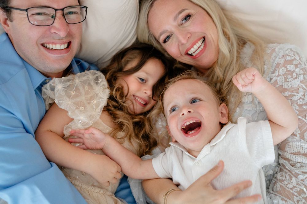 A family laughs together while cuddling on white bedding in natural light.