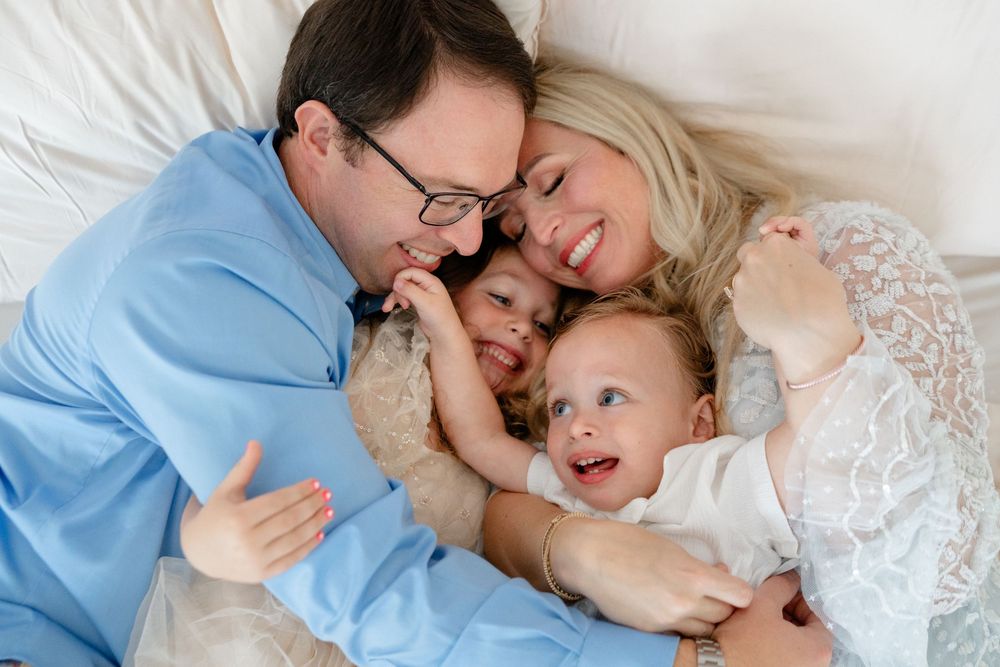 Family sharing intimate cuddles and laughter while relaxing together on white bedding.