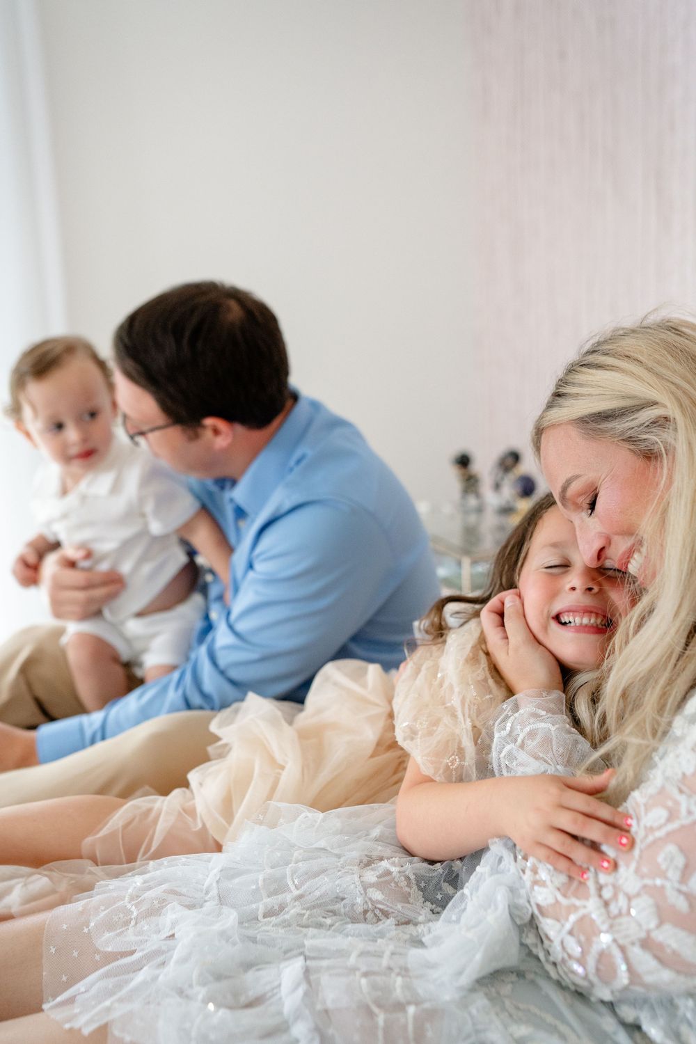 A family shares joyful moments together while lying down in white and lace outfits against a light background.