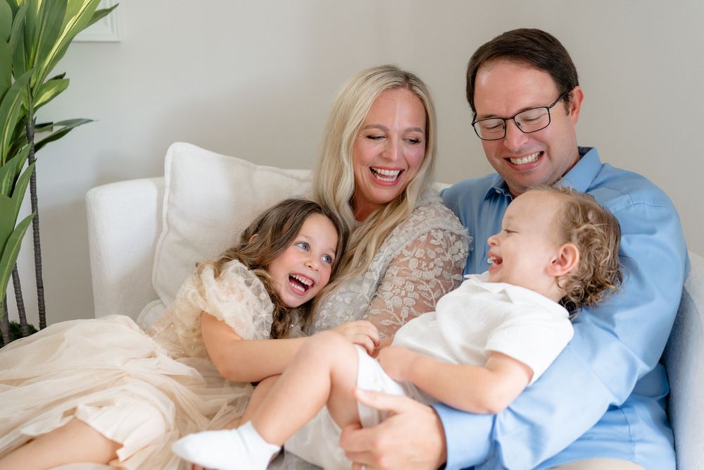 A happy family lounges together on a white sofa with indoor plants and blue decor in the background.
