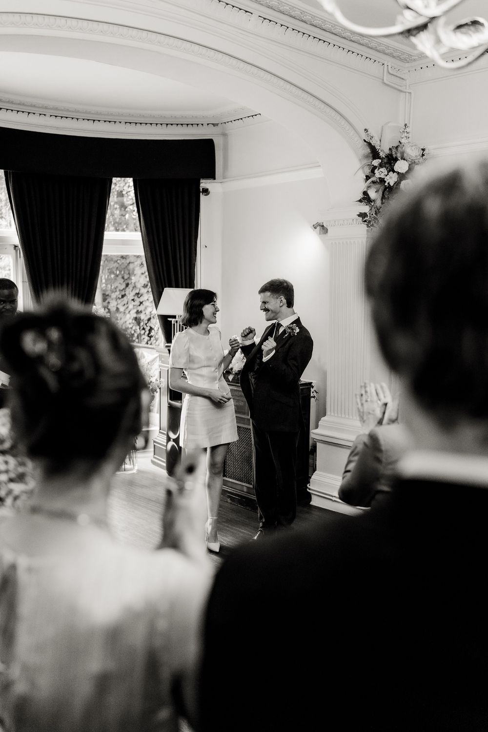 A black and white photo of a wedding ceremony in an elegant room with dark curtains and guests watching.