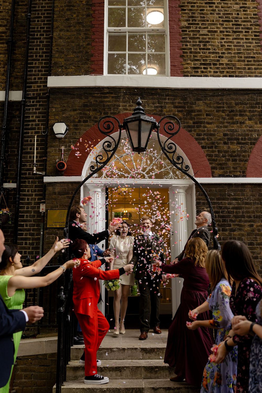 A group of people throw confetti outside a brick building entrance with ornate iron archways during a celebration.