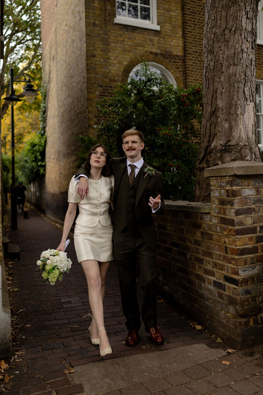 A newlywed couple walks along a brick path beside an old building with autumn foliage in the background.