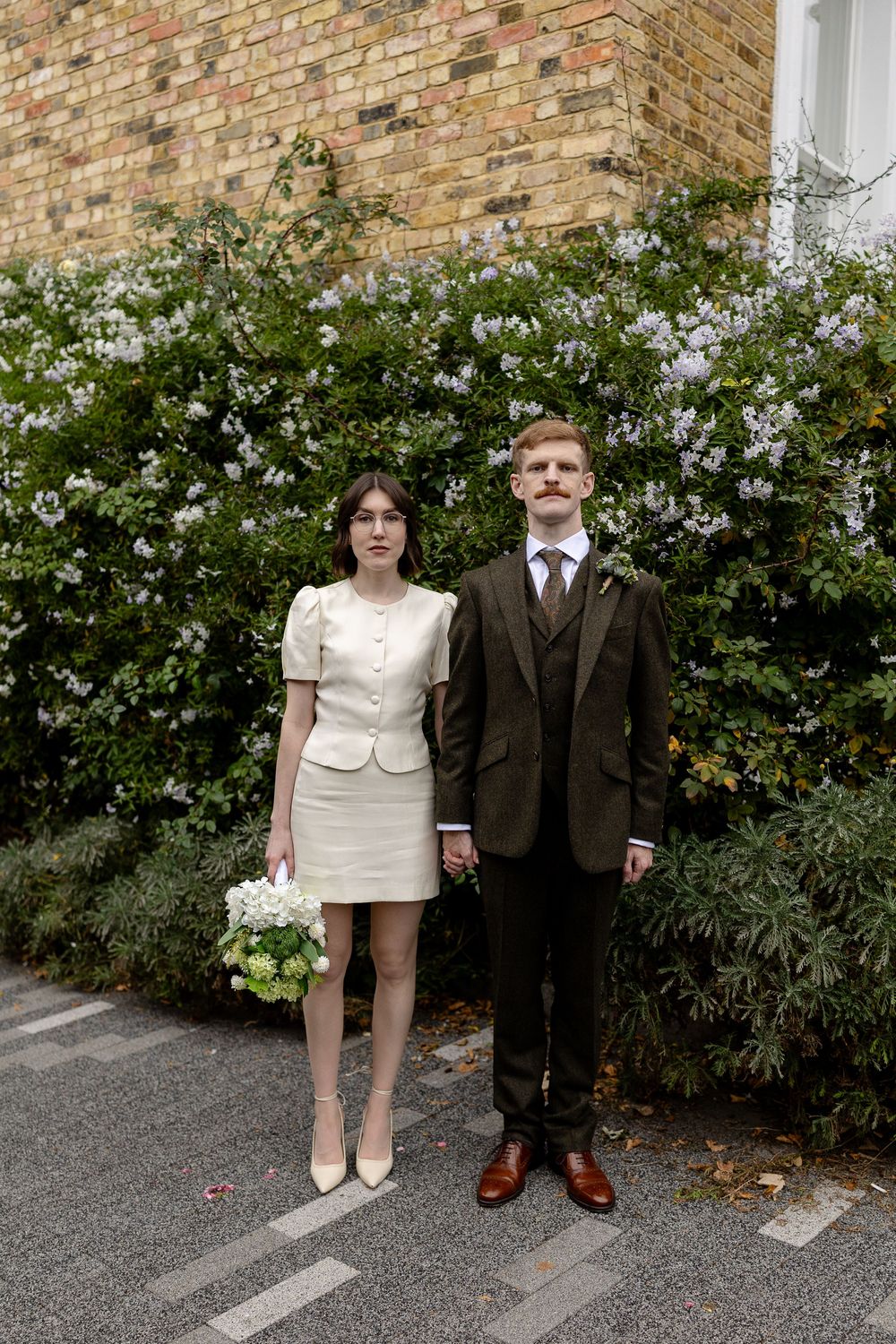 A couple in formal attire stands against a brick wall with flowering bushes in the background.