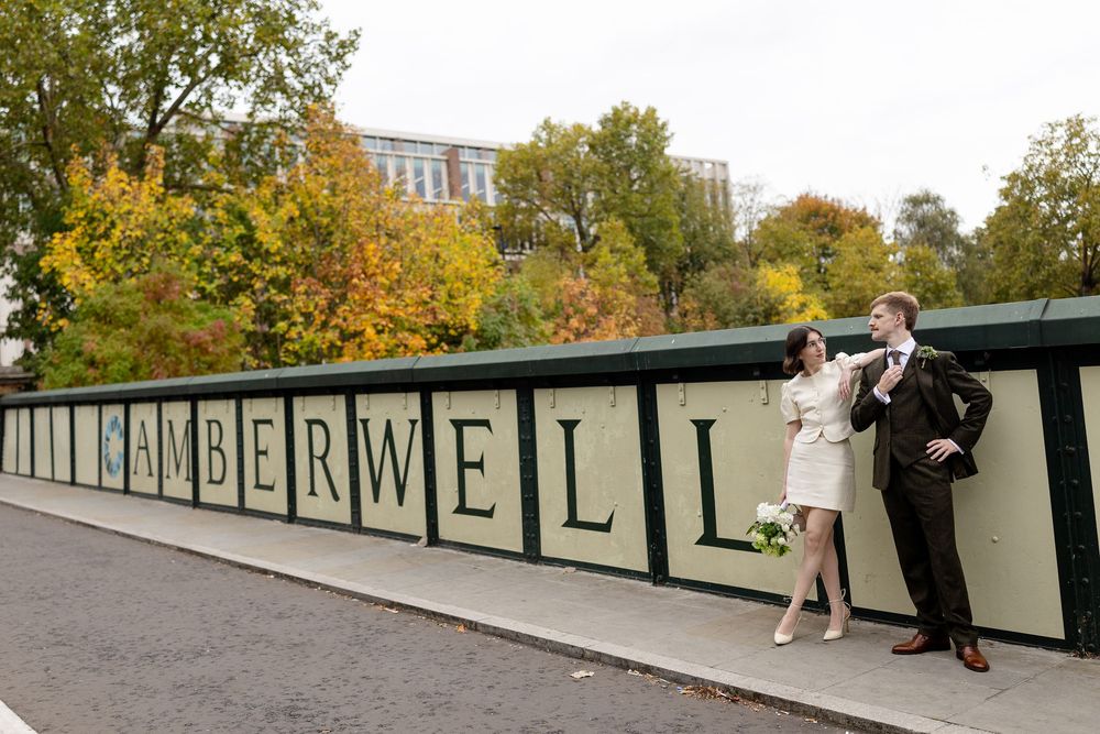 A couple poses in front of a wall with 'Camberwell' text during autumn with yellow trees in the background.