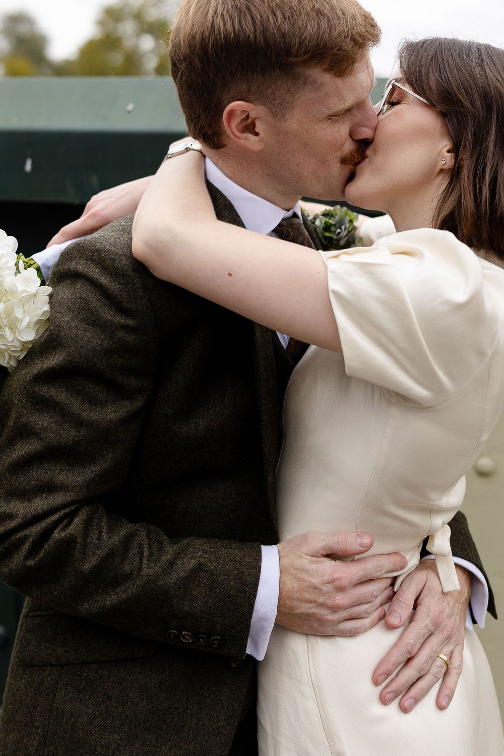 A romantic wedding kiss captured outdoors with the bride in cream dress and groom in dark suit.