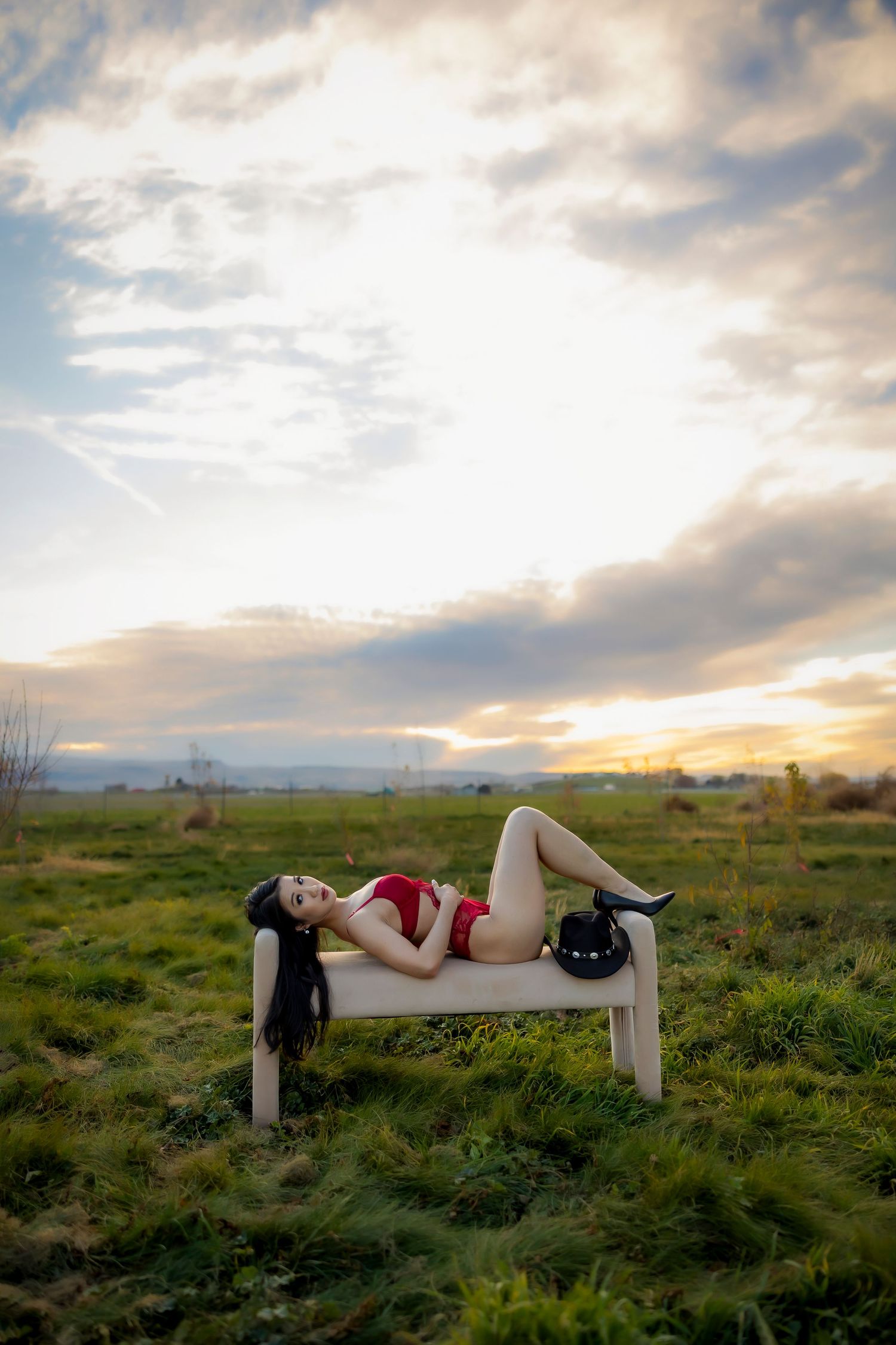 Dark haired woman wearing red lingerie lying on bench with black heels and black cowboy hat in grassy area